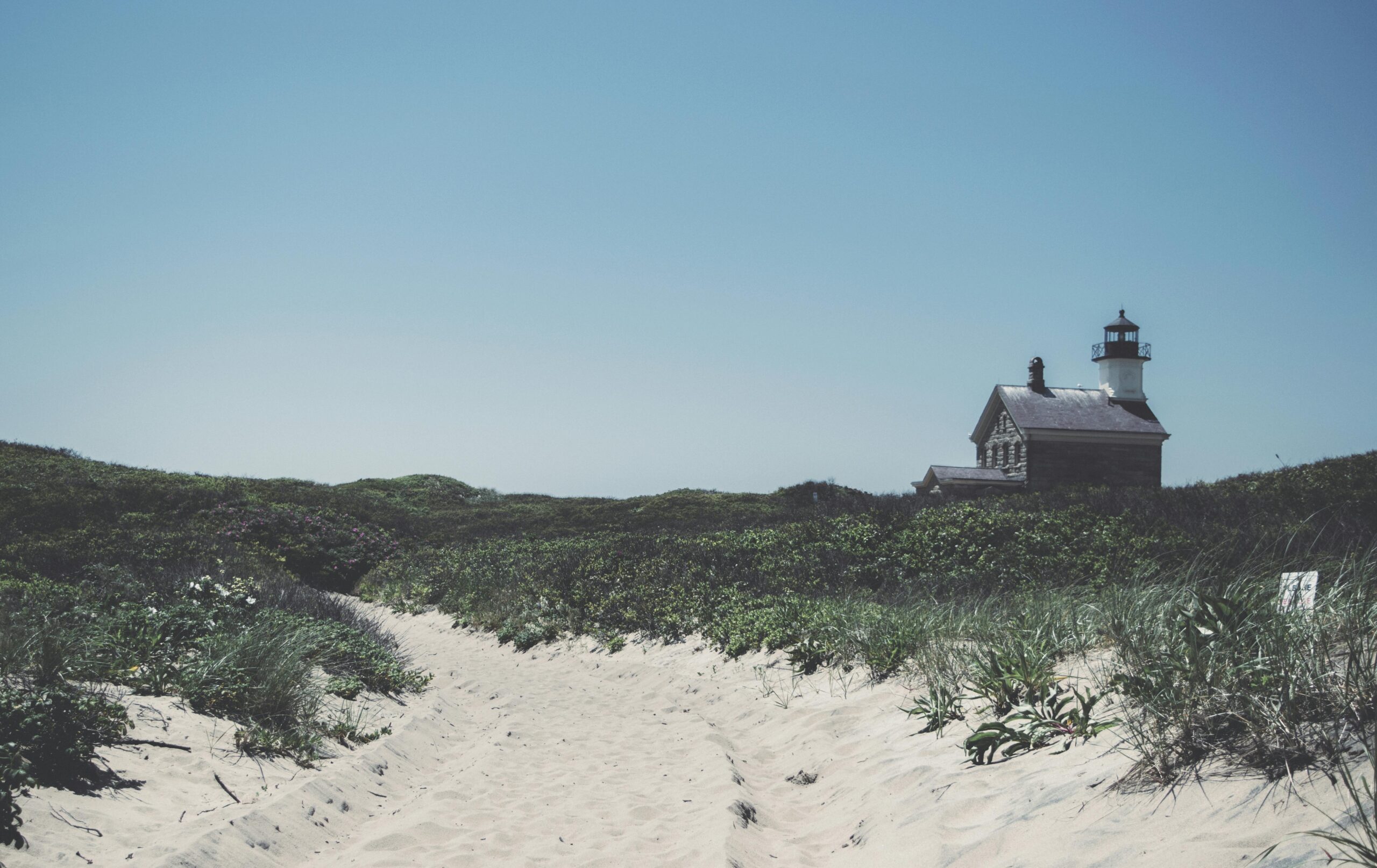 Scenic view of Block Island Lighthouse with sandy path and lush green dunes under blue sky.